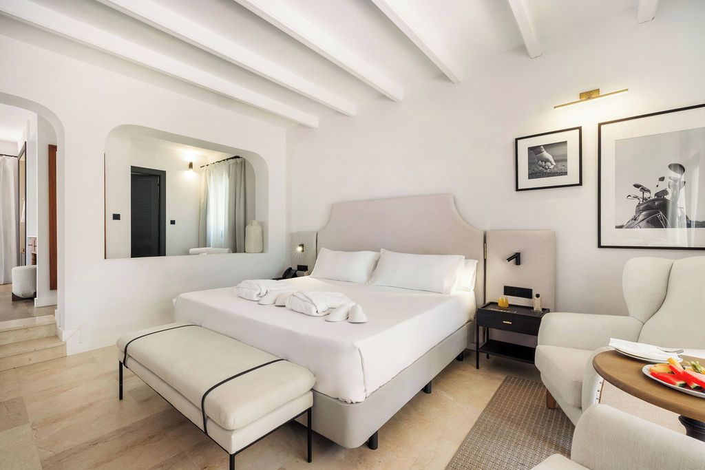 Habitación de hotel con cama, reposabancos, mesita de noche, silla y una pequeña mesa con un plato de fruta.
