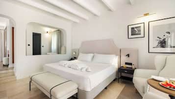Habitación de hotel con cama, reposabancos, mesita de noche, silla y una pequeña mesa con un plato de fruta.