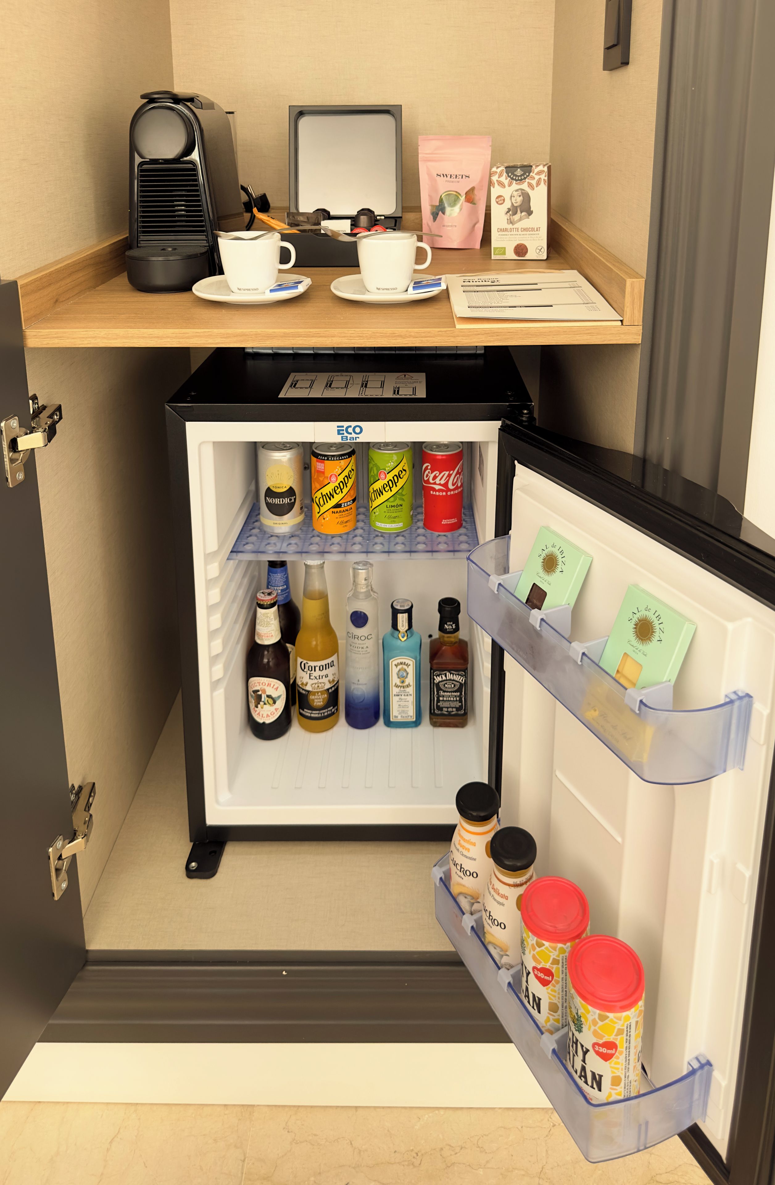 A minibar with a coffee machine, two cups, a chocolate bar, and various beverages.