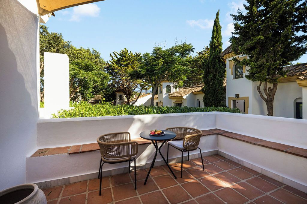 A terrace with two wicker chairs and a small table, overlooking a courtyard with greenery and buildings.