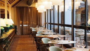 L’intérieur d’un restaurant, avec des tables dressées pour le service, de grandes fenêtres et des lampes suspendues.