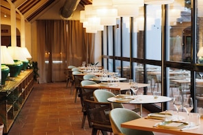 Interior de restaurant amb taules preparades per dinar, grans finestres i lluminàries penjants.
