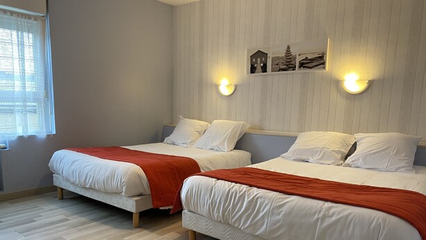 Une chambre avec deux lits, chacun recouvert de draps blancs et d’une couverture rouge. On y trouve des lampes fixées au mur et une fenêtre ornée de rideaux à motifs.