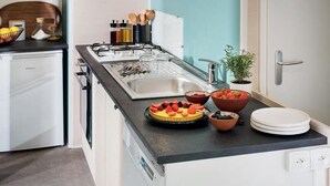 A compact kitchen with a gas stove, sink, and a small refrigerator. There are bowls of fruit on the counter and a potted plant near the sink.