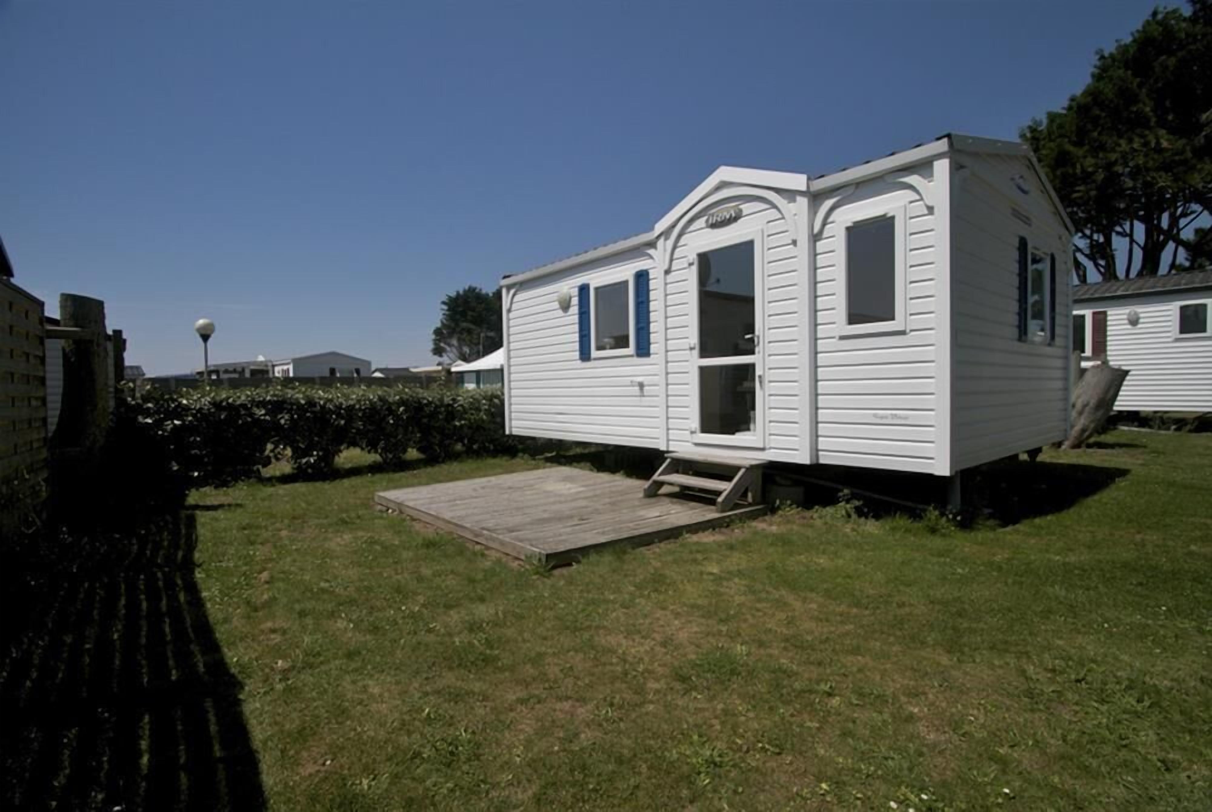 A white mobile home with a small porch and a ramp, surrounded by grass and trees.