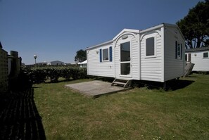 A white mobile home with a small porch and a ramp, surrounded by grass and trees.