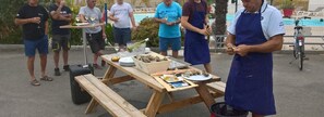 Un groupe de personnes est réuni autour d’une table de pique-nique en bois, avec de la nourriture et des boissons, à l’extérieur, près d’une piscine.