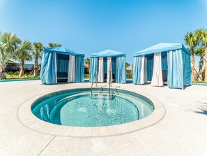 Piscina com um elemento central de água, rodeada por toldos azuis e palmeiras.