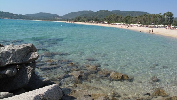 A beach with clear water, rocky foreground, and a sandy shore with umbrellas and people.