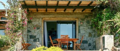 Un edificio de piedra con una pérgola de madera, una mesa y sillas afuera, y un banco de piedra.