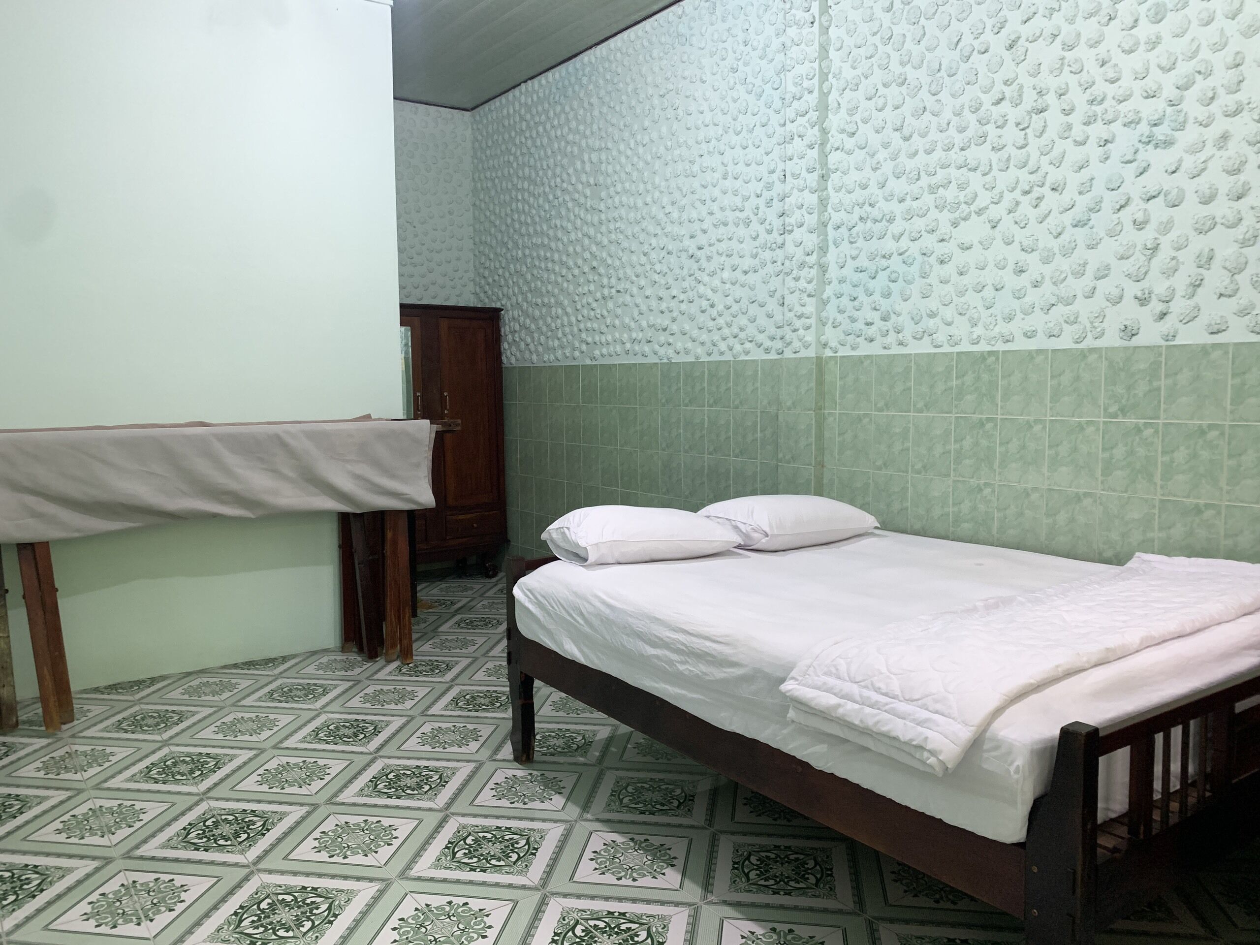 A small, clean room with a single bed, a wooden wardrobe, and a tiled floor.
