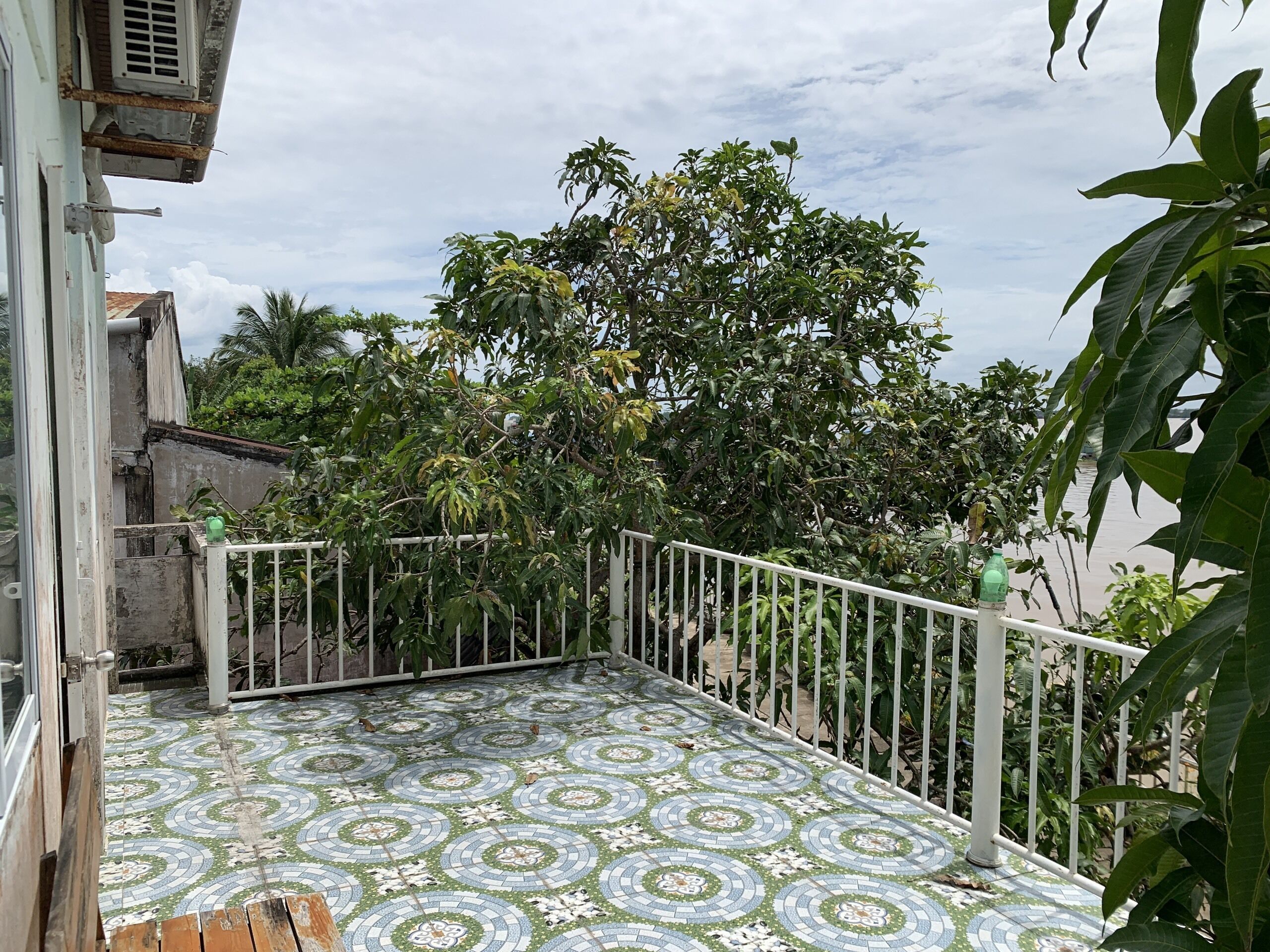 Un balcone piastrellato con ringhiera in metallo, immerso nel verde e con cielo sereno.