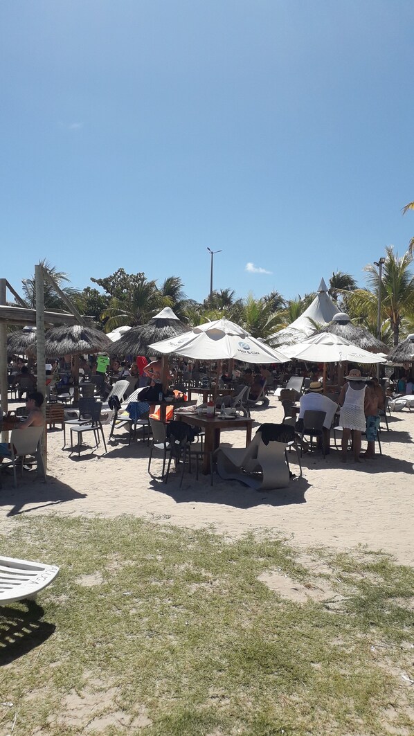 Um restaurante à beira-mar com mesas ao ar livre, palmeiras e toldos de palha.