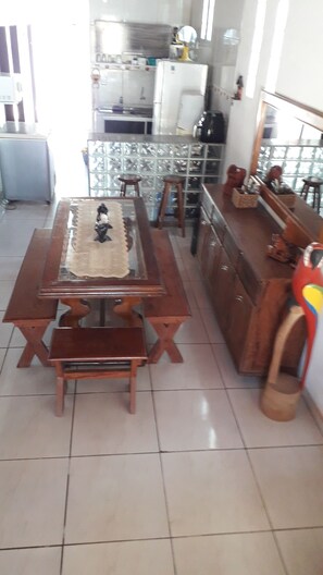 Uma área de refeições com mesa e bancos de madeira, cozinha ao fundo e vista para o exterior por uma porta de vidro.