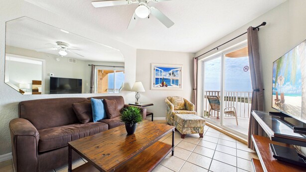 A living room with a brown leather sofa, a wooden coffee table, a patterned armchair, a TV, and a balcony with a view of the ocean. - Surf Beach 503 - Spectacular Sunsets - Beach Front (Treasure Island)