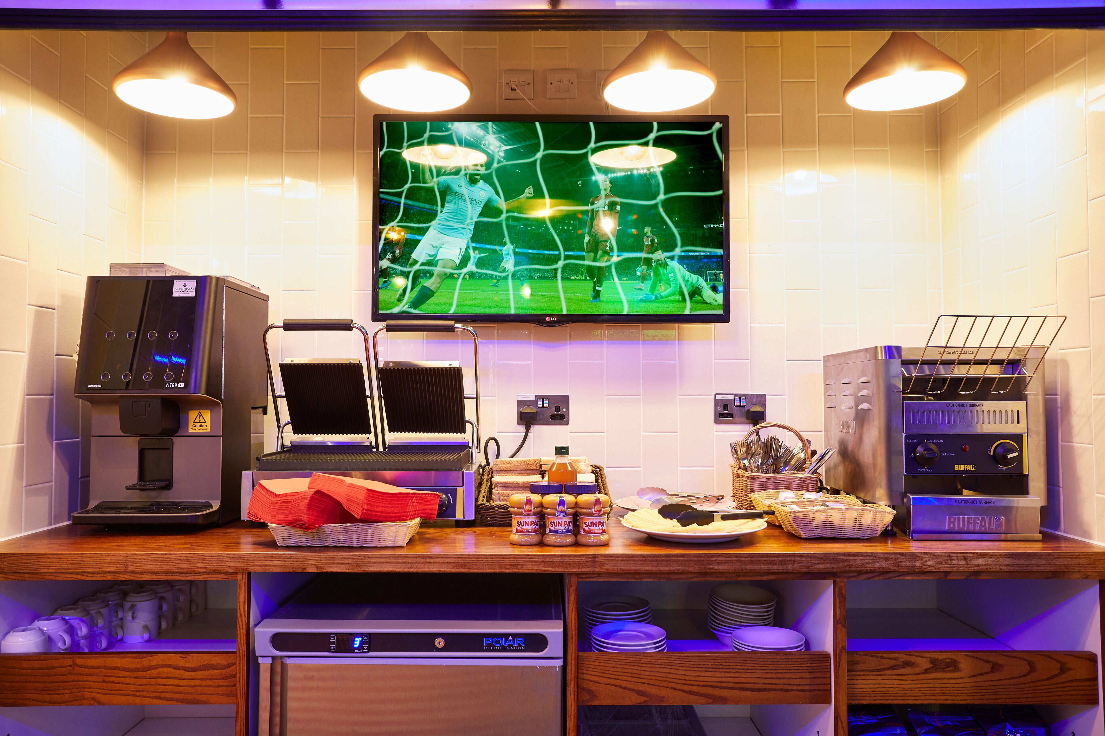 A kitchen area with a coffee machine, a microwave, a toaster, and a waffle maker. There is a television mounted on the wall displaying a soccer game.