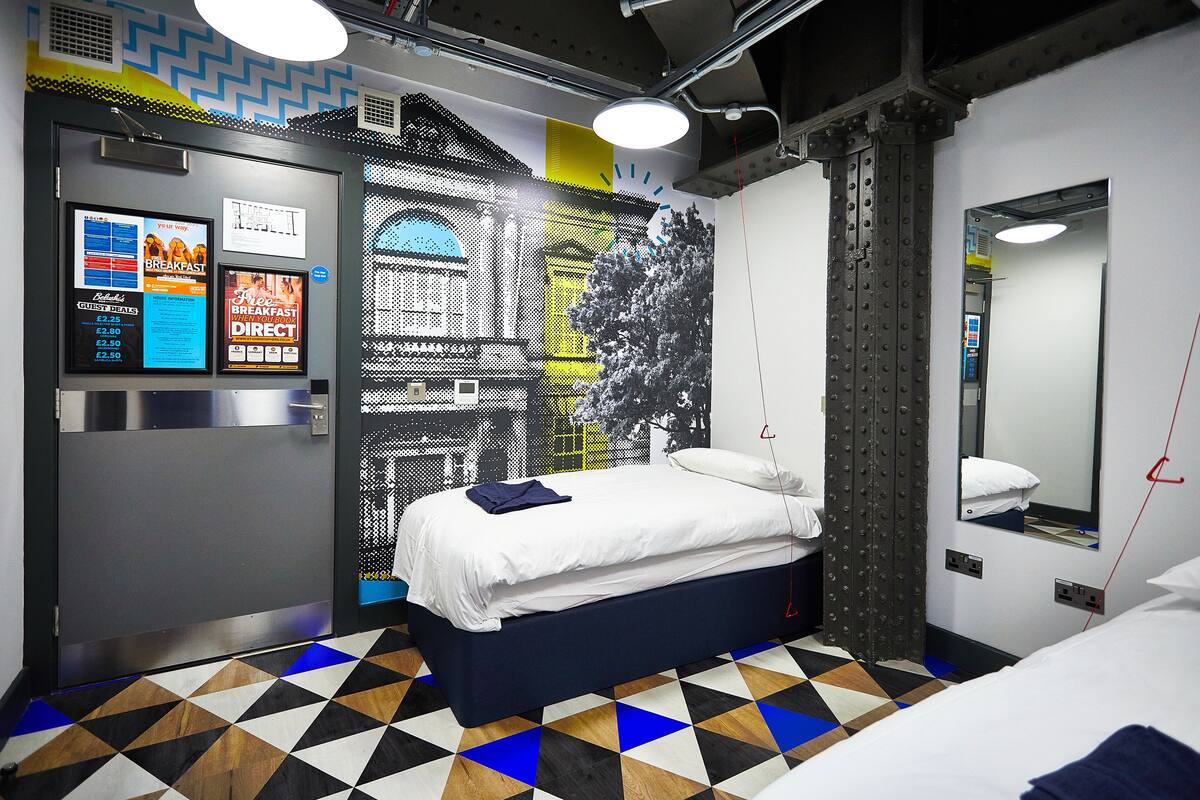 Habitación de hotel con cama, un panel digital de menú, piso de estampado geométrico y un gran mural en la pared.