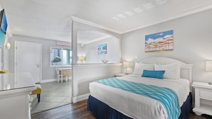 Ein Hotelzimmer mit einem Bett, Nachttischen, einem Schreibtisch und einem Sessel. Es gibt ein Fenster mit Jalousien und eine Tür.