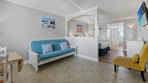 Ein modernes Wohnzimmer mit einem blauen Sofa, einem gelben Sessel und einem weißen Couchtisch. Es gibt eine Küchenzeile mit einer weißen Arbeitsplatte und einem Esstisch.