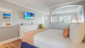 Une chambre d’hôtel avec un lit, une télévision, une commode, une chaise, une fenêtre donnant sur des immeubles et un tableau accroché au mur.