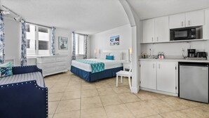 A compact room with a bed, a small desk, a chair, a kitchenette with a microwave and refrigerator, and a window with curtains. - Pelican Pointe 417:Walk to Beach~Pool~Free Parking (Clearwater Beach)