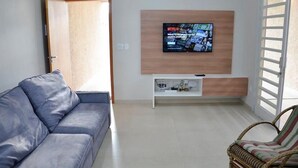A living room with a grey sofa, a wicker chair, a TV mounted on a wooden wall, and a sliding glass door. - (Olímpia)