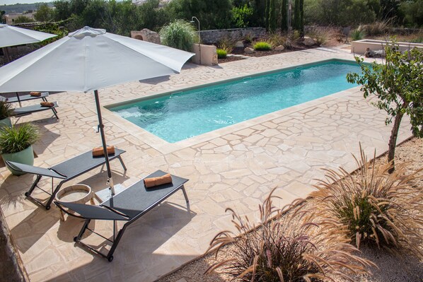 Una piscina rectangular con baldosas de piedra, rodeada de sillones de descanso y sombrillas, con plantas en macetas y un sendero de piedra.
