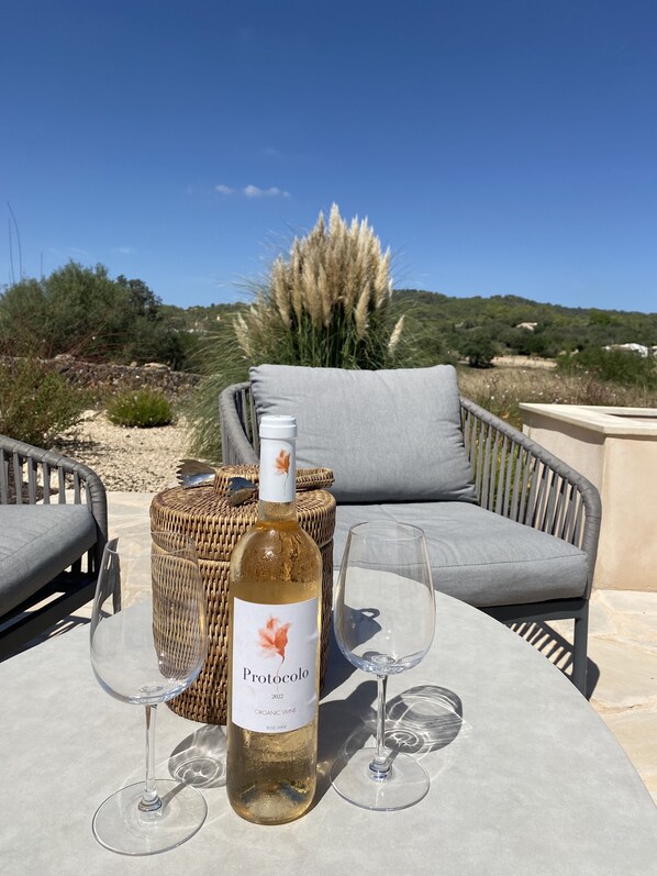 A table with a bottle of wine and two glasses, set on an outdoor patio with wicker furniture and a view of a hill and vegetation.