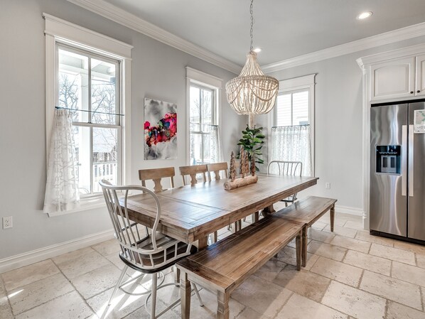 A dining area with a wooden table, chairs, and a bench. There is a chandelier above the table, and a refrigerator is visible to the right.