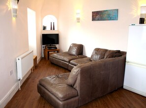 A living room with a brown leather sofa, a flat-screen TV, a small wooden side table, and a refrigerator. - Cotswolds Valley Accommodation-Excl Prop (Stroud)