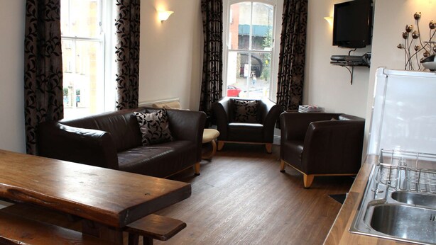 A modern living room with a wooden dining table, two sofas, a TV, and a kitchen area with a sink. - Cotswolds Valley Accommodation-Excl Prop (Stroud)