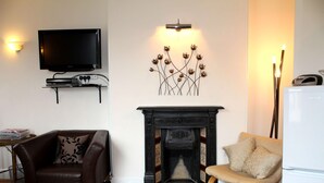 A living room with a fireplace, a television mounted on the wall, and two chairs. - Cotswolds Valley Accommodation-Excl Prop (Stroud)