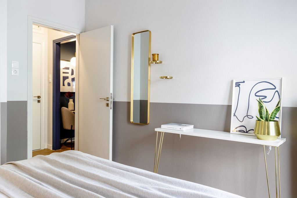 A hotel room with a bed, a bedside table with a mirror, a small shelf with a plant, and a door leading to a bathroom.