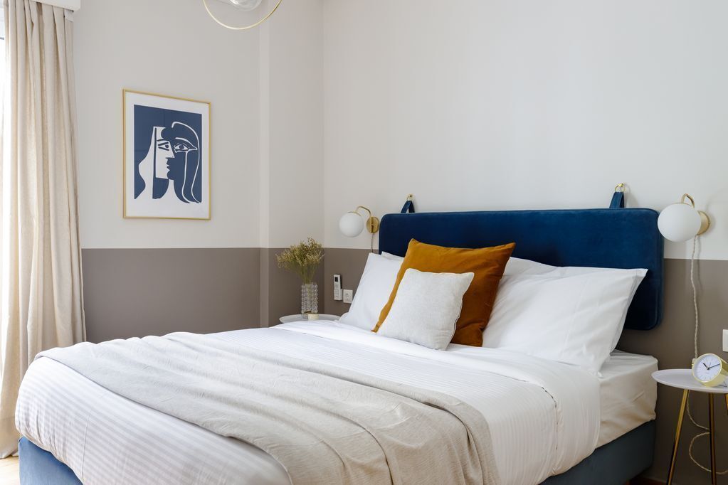 Un lit bien fait, avec une tête de lit bleue, du linge de lit blanc et un coussin de couleur moutarde. On aperçoit une petite table de chevet avec une lampe et une horloge. Une œuvre d’art encadrée est visible au mur.