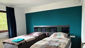 A bedroom with two beds, a window with curtains, and a teal-colored wall. - 3 Room Appartment (70 m2) with 2 Bedrooms, Balcony & Garage (Neckarsulm)