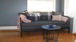 A grey sofa with patterned cushions, a small round table, and a window with blinds.