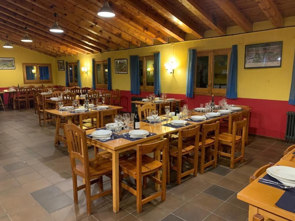 Un comedor con varias mesas y sillas de madera, dispuesto en una habitación amplia con techo de madera y suelo de baldosas.
