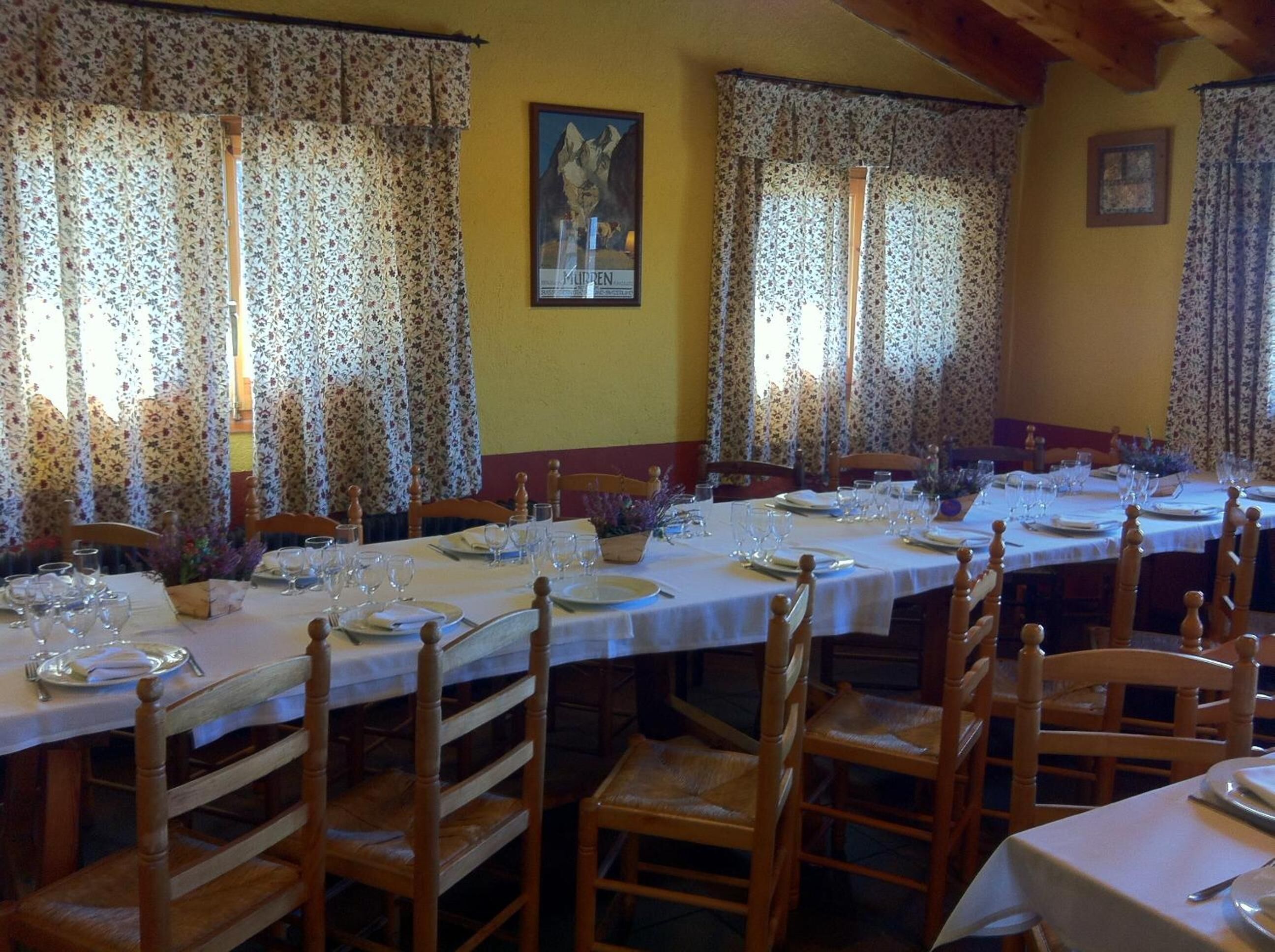 Un comedor con varias mesas dispuestas para una comida, sillas de madera y cortinas con motivos florales.