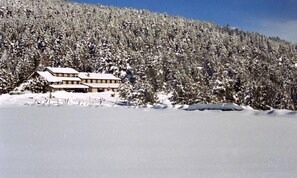 A snow-covered lodge with a symmetrical facade and a flat roof, surrounded by dense forest. - Refugi Cap del Rec - Hostel (Lles)