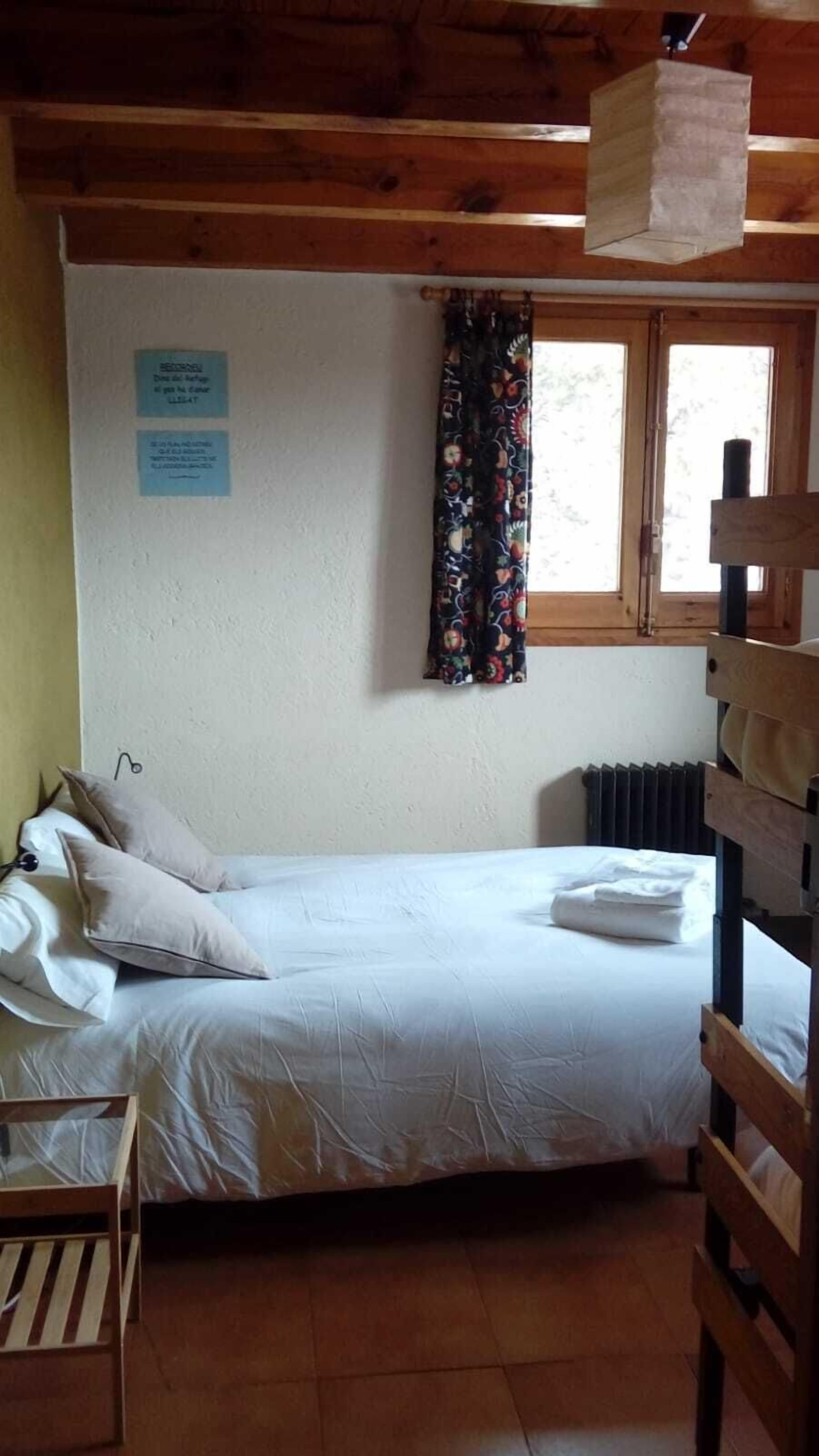 Una habitación pequeña y acogedora con una cama individual, una cama nido de madera a la derecha, una ventana con cortinas florales y un letrero en la pared.
