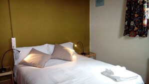 A neatly made bed with white linens and beige pillows, a bedside table with a lamp, and a wall with a floral tapestry. - Refugi Cap del Rec - Hostel (Lles)