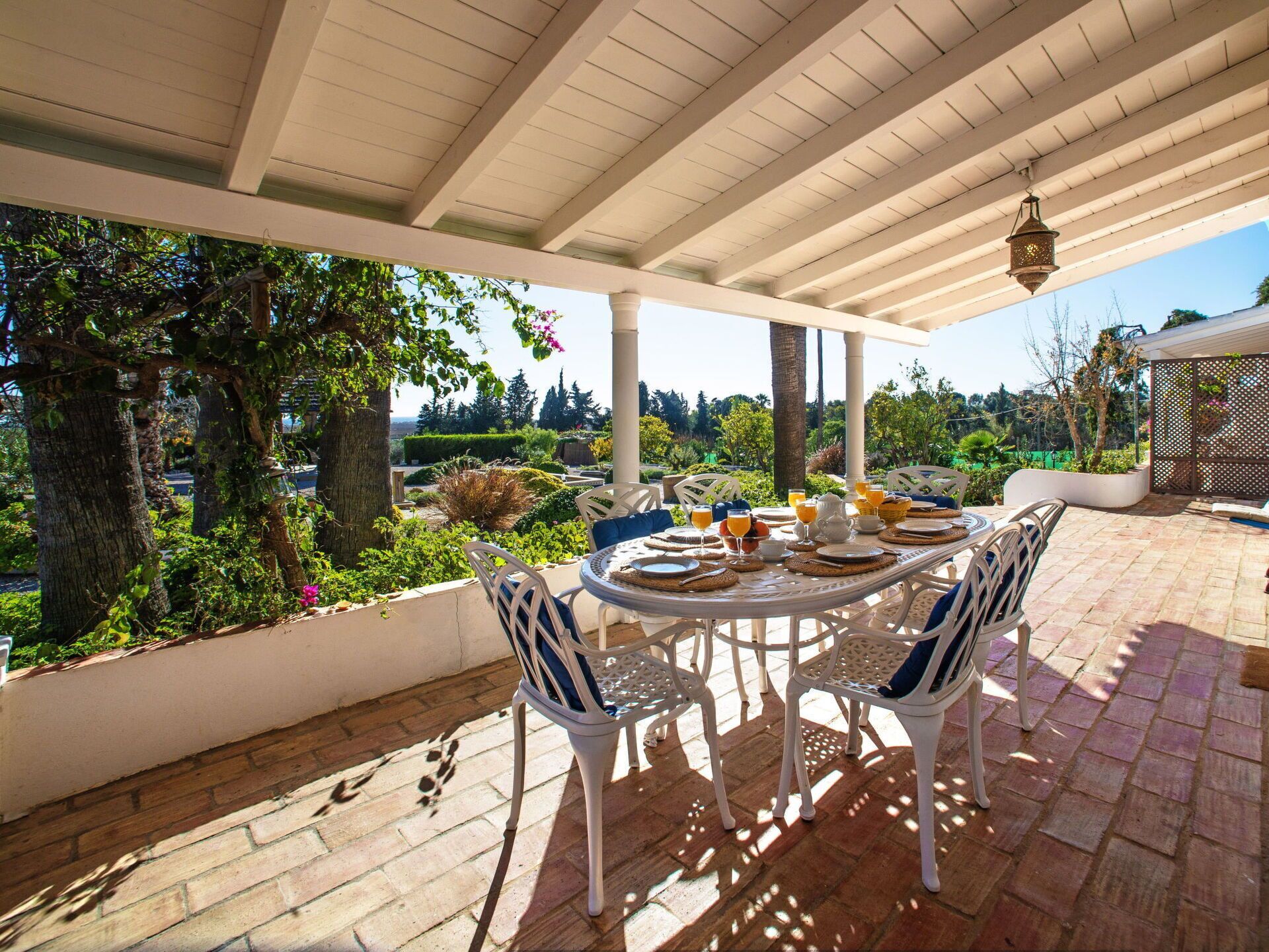 Un patio al aire libre con una mesa redonda preparada para comer, rodeada de sillas blancas, bajo una zona cubierta con vistas a un jardín exuberante y árboles.