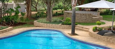 Una piscina con borde de azulejos, rodeada de un jardín ajardinado con árboles y un muro de piedra.