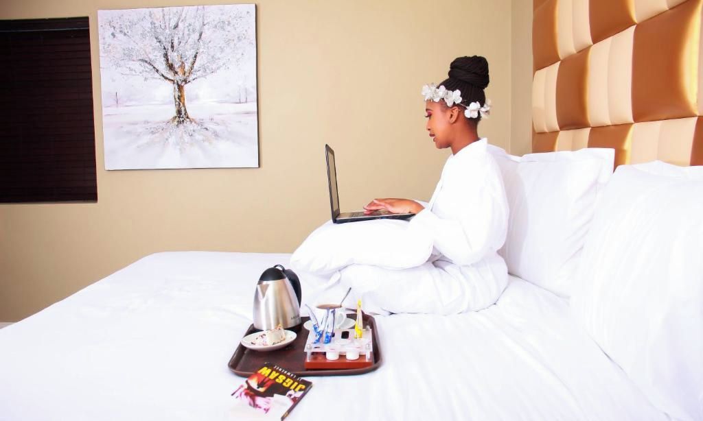 Uma pessoa sentada em uma cama com um laptop, uma bandeja com uma xícara, um prato de comida e uma revista.