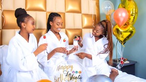 Three individuals in white robes sitting on a bed with a 'Happy La Fiesta' sign, a tufted headboard, and balloons. - La Fiato Luxury Lodge (Polokwane)