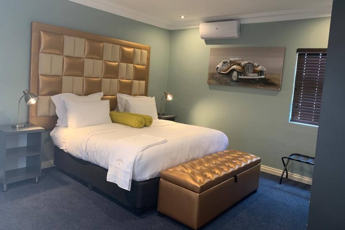 Een slaapkamer met een groot bed, een hoofdbord met een gestoffeerde uitstraling, een nachtkastje met een lamp, een bankje aan het voeteneinde van het bed, en een wandairconditioner.