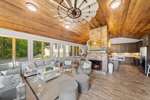 Relax in this spacious living area with a high ceiling, featuring a circular beam design, a fireplace, and large windows offering views of the surrounding greenery.