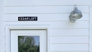 Una puerta blanca con un letrero de Cedar Loft arriba, una ventana con vista a árboles, y una lámpara fijada a la pared.