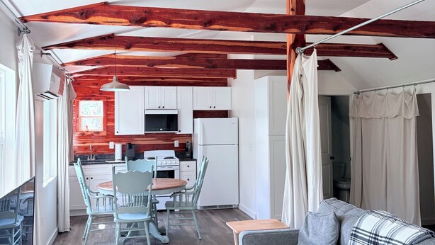 Un interior acogedor con vigas de madera a la vista, una zona de comedor con mesa y sillas, una kitchenette y un sofá con un cojín de cuadros.