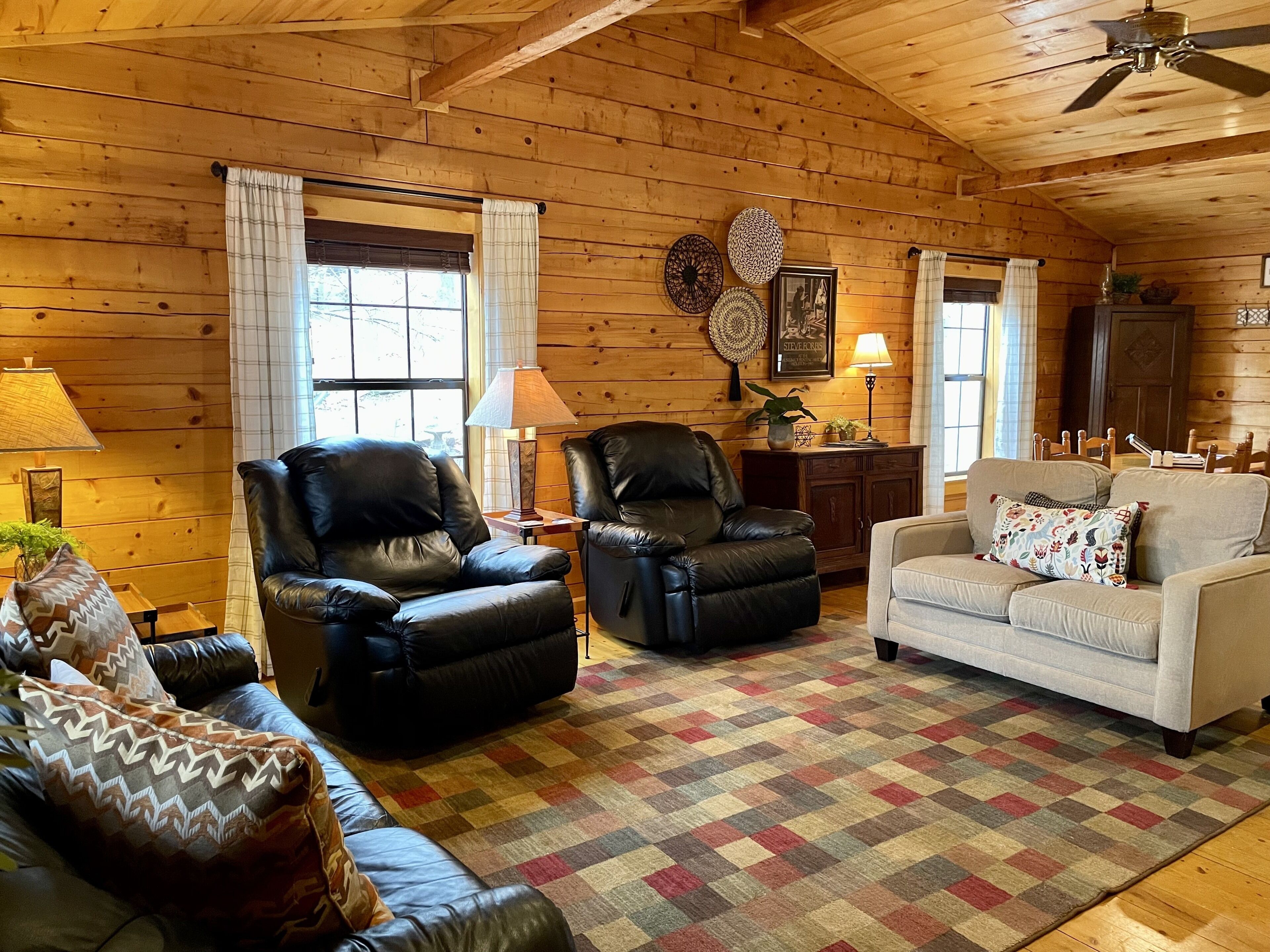 A cozy cabin interior with wooden walls, two leather chairs, a white sofa, and a patterned rug.
